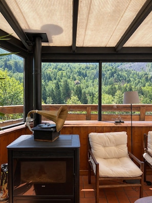 Ein gemütlicher Außenbereich mit Holzofen, einem Tisch und einem Grammophon, mit Blick auf eine bewaldete Landschaft.
