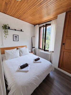 A bedroom with a wooden ceiling, a large bed with white bedding, a radiator, and a window with curtains. - Hotel Au Bon Accueil (Les Deux Alpes)