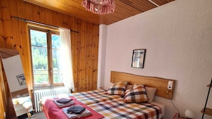 A bedroom with a wooden ceiling, a large bed with a checkered bedspread, a bedside table with a lamp, and a window with a view of greenery. - Hotel Au Bon Accueil (Les Deux Alpes)