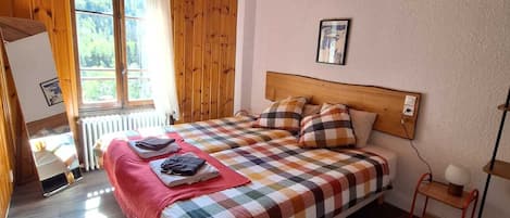 Un dormitorio con techo de madera, una cama grande con colcha a cuadros, una mesita de noche con lámpara y una ventana con vistas a zonas verdes.