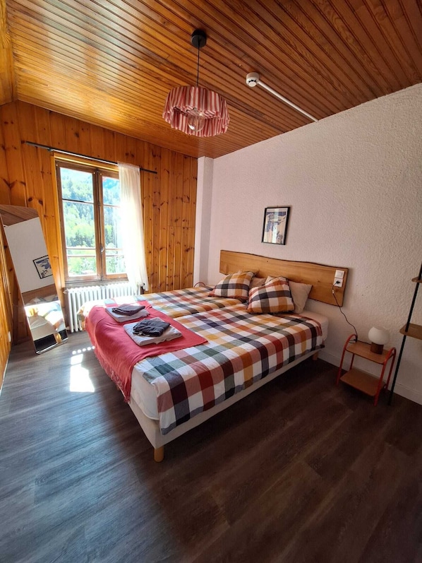 A bedroom with a wooden ceiling, a large bed with a checkered bedspread, a bedside table with a lamp, and a window with a view of greenery. - Hotel Au Bon Accueil (Les Deux Alpes)