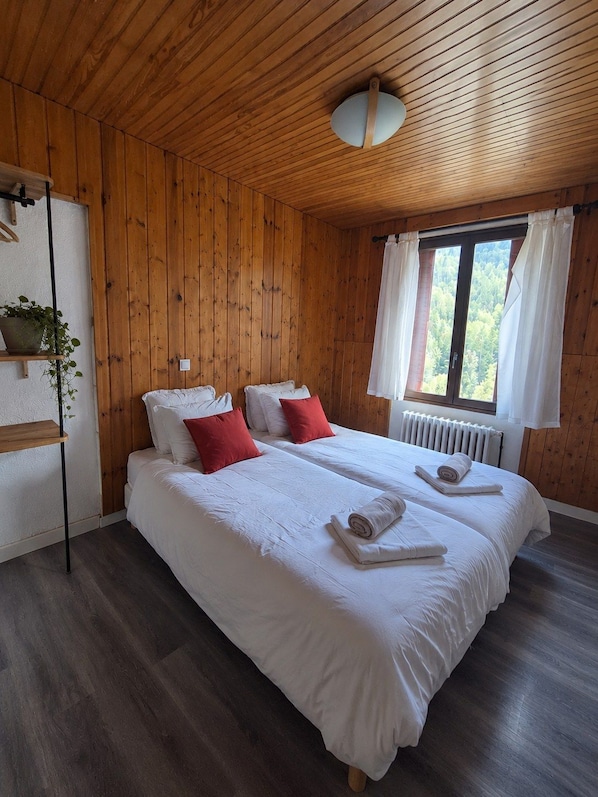 A double bed with white bedding and red pillows, a wooden ceiling, a window with white curtains, and a radiator. - Hotel Au Bon Accueil (Les Deux Alpes)