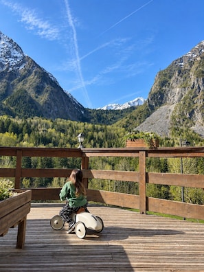 A wooden deck with a person in a wheelchair, overlooking a mountainous landscape with snow-capped peaks and dense forest. - Hotel Au Bon Accueil (Les Deux Alpes)