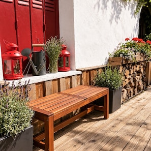 A wooden bench on a wooden deck with a white wall and red shutters in the background. - Hotel Au Bon Accueil (Les Deux Alpes)