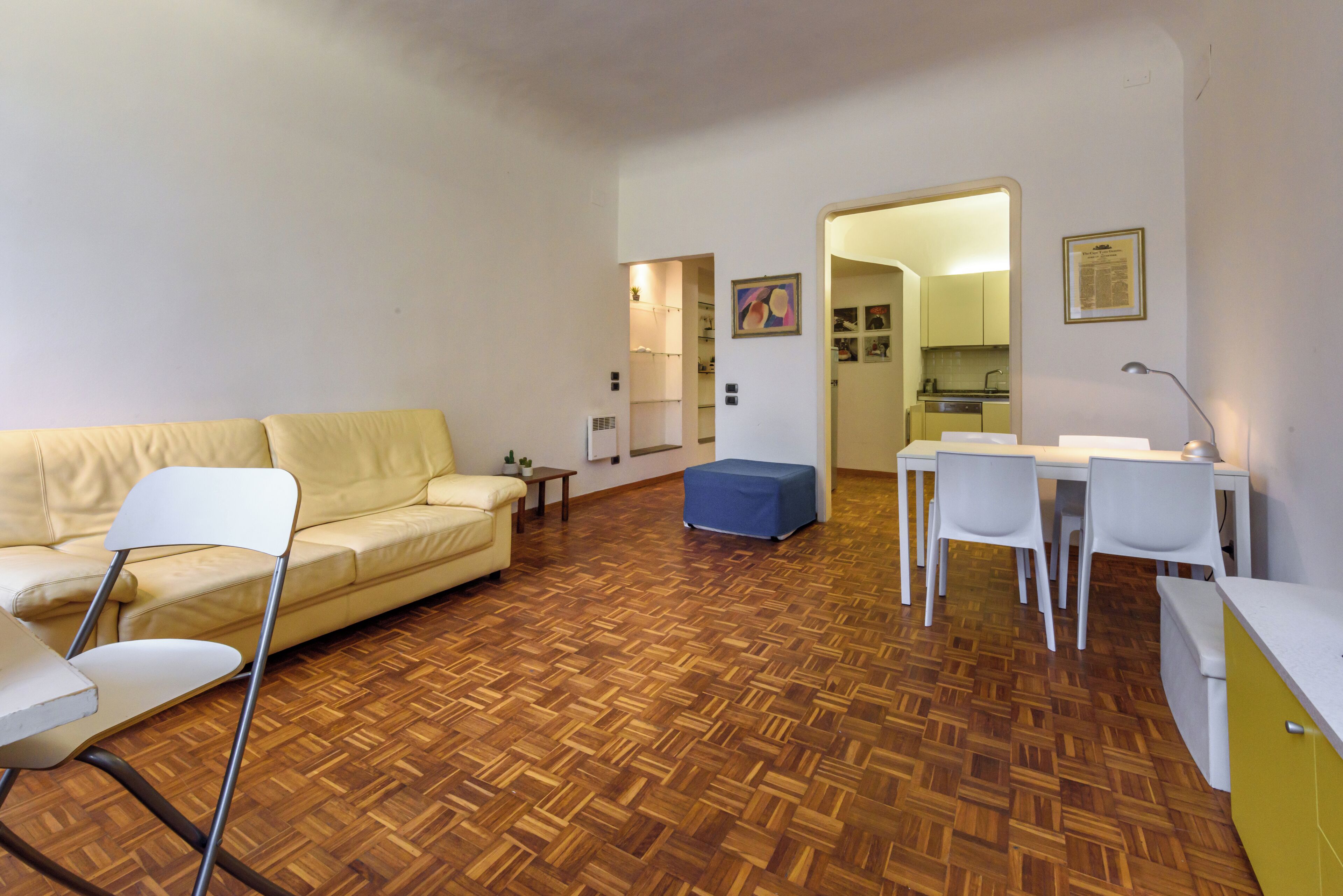 A living room with a wooden parquet floor, a yellow sofa, a white chair, a small table, and a blue ottoman.