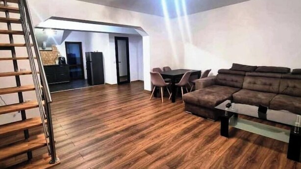 Ein großzügiger Wohnbereich mit Holzboden, einem braunen Sofa, einem Esstisch mit Stühlen und einer Küche im Hintergrund.