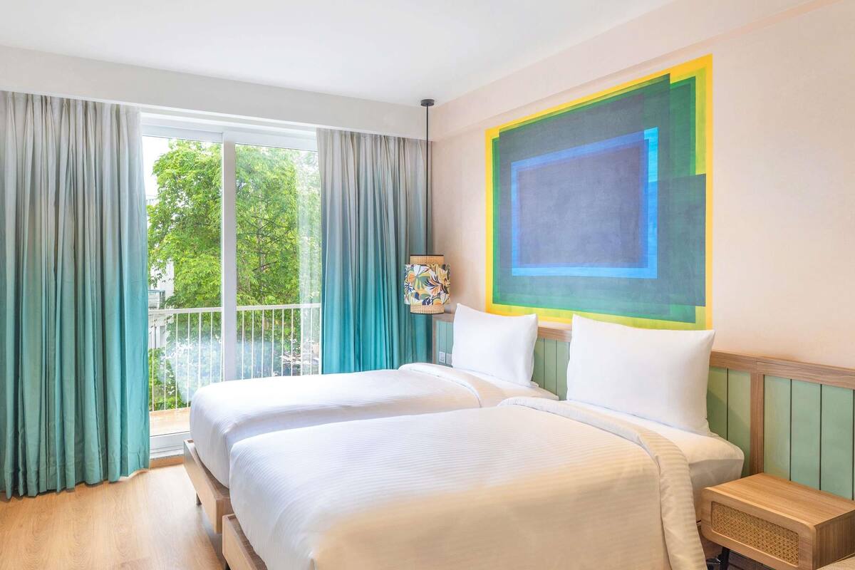 Habitación de hotel con dos camas, un ventanal con cortinas, un cuadro abstracto de colores y una mesita de noche de madera.