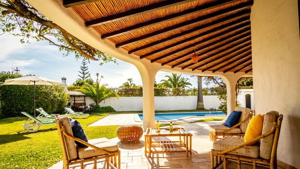 Une terrasse extérieure aménagée avec des meubles en osier, une piscine et un espace couvert doté d’un plafond en bois.
