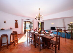 A dining area with a large wooden table set for six, surrounded by wooden chairs. There is a bar area with stools, a television, and a fireplace. - Villa with heatable pool and BBQ in Albufeira (Albufeira)