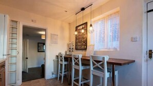 Une cuisine avec une table en bois, des chaises blanches, un luminaire suspendu et une fenêtre avec des rideaux.