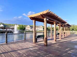Una terraza de madera con un pérgola, un banco y vista a una casa al otro lado del agua.