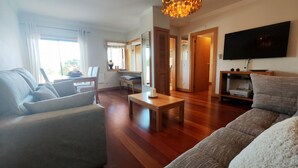 Un salon moderne avec un canapé, une table basse et un téléviseur. Il y a également un coin repas avec une table et des chaises. La pièce est dotée d’un parquet et d’une grande fenêtre.