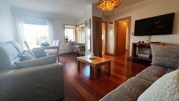 Una sala de estar moderna con un sofá, una mesa de centro y un televisor. Cuenta con una zona de comedor con una mesa y sillas. El piso es de madera y hay un ventanal grande.