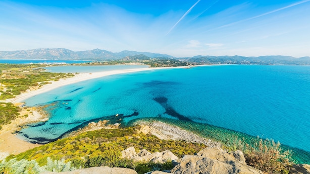 Un paysage côtier avec une eau turquoise cristalline, une plage de sable et des montagnes à l’horizon.