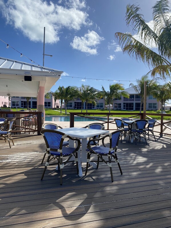 An outdoor deck with tables and chairs, a pool, and a building with a white facade in the background. - Fabulous beachfront condo (Treasure Cay)