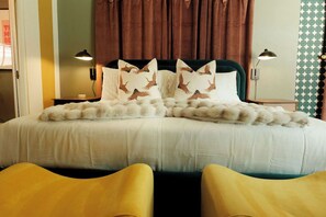 Un lit avec une literie blanche, un plaid blanc moelleux et des coussins décoratifs ornés de motifs d’oiseaux. Il y a deux lampes de chevet et un rideau.