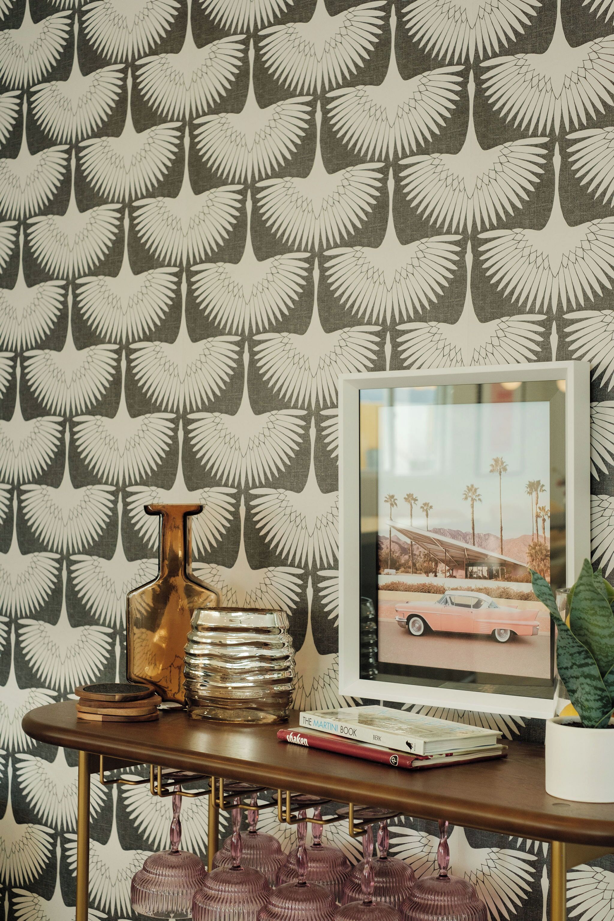 A room with a patterned wallpaper, a framed picture on the wall, a table with a book, a vase, and a plant.