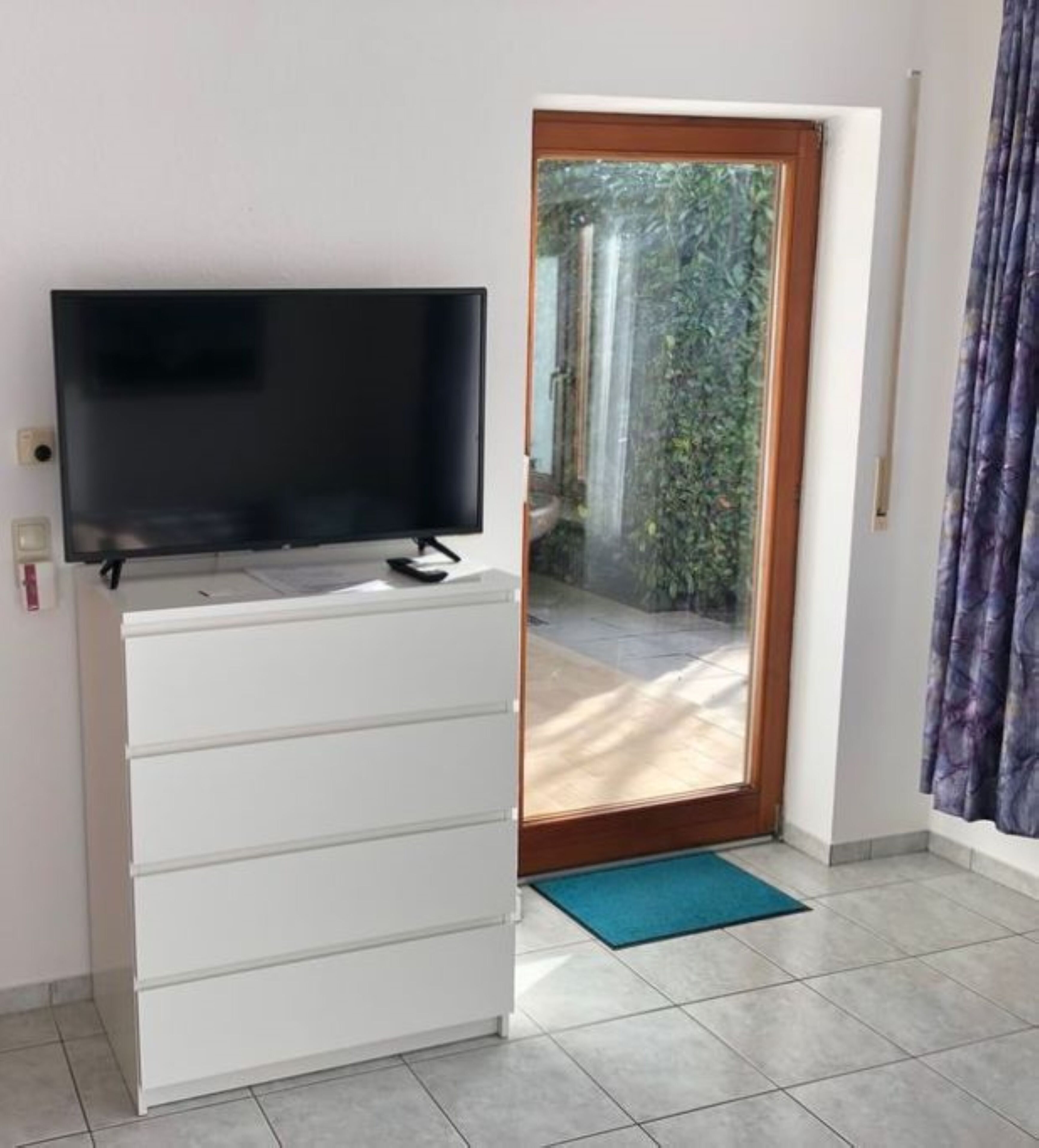 Een kamer met een flatscreen televisie op een witte kast, een schuifdeur naar een terras en een blauwe mat op de vloer.