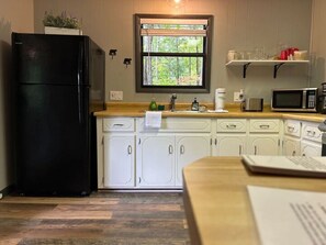 Una cocina con armarios blancos, un refrigerador negro, una encimera de madera y una ventana con vistas a árboles, además de un microondas.