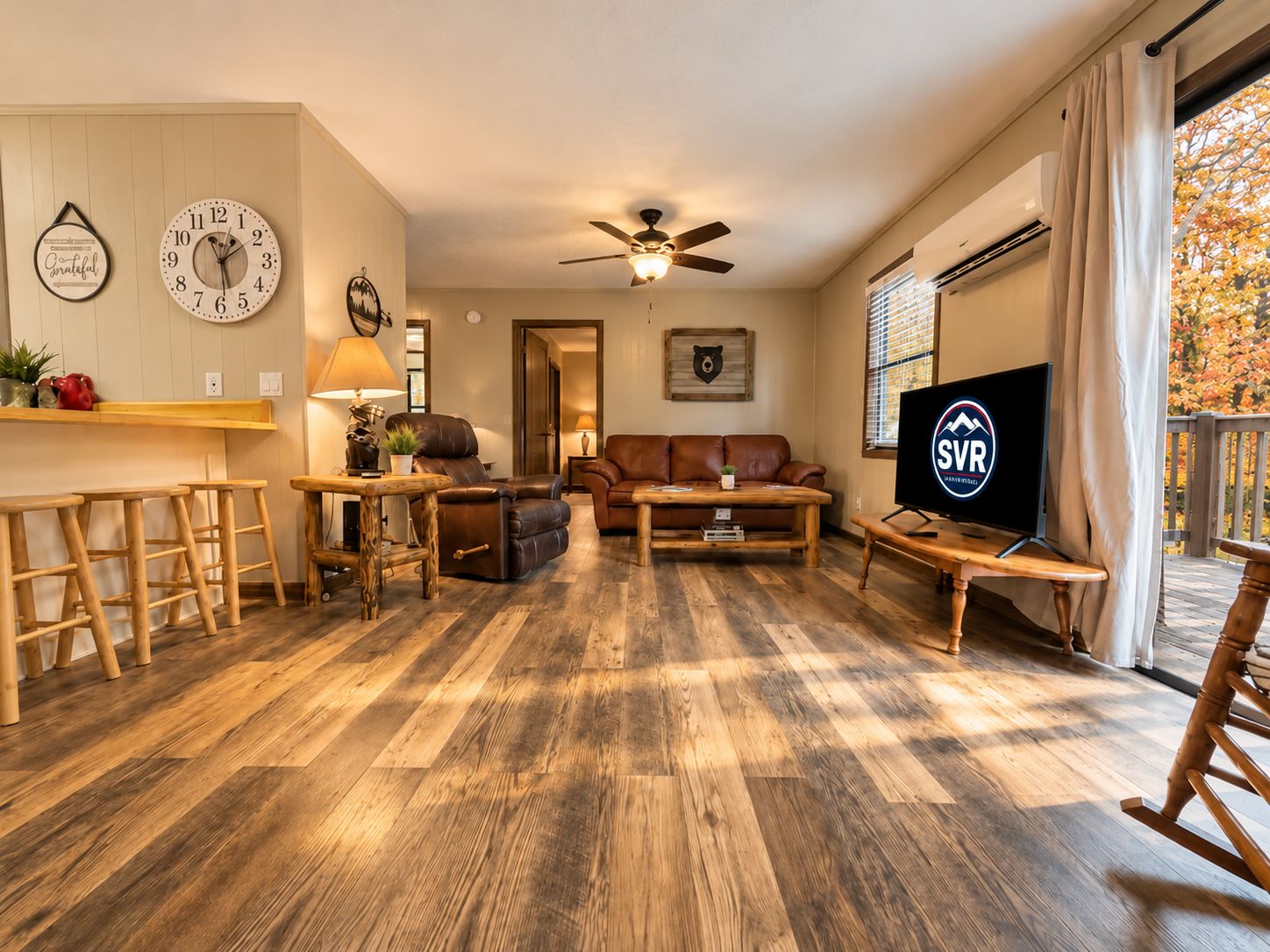 A living room with a sofa, coffee table, and TV. There are wooden floors, a ceiling fan, and a window with blinds.