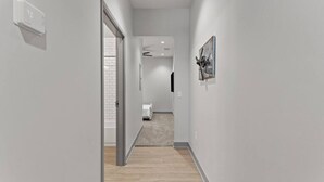 A narrow hallway with a wooden floor, white walls, and a door leading to a room with a white bed and a brick wall. - The Gatsby Getaway, Downtown LEX (Lexington)