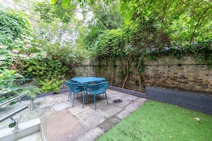 Un patio con mesa y sillas, rodeado de abundante vegetación y un muro de ladrillo.