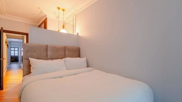 Ein ordentlich bezogenes Bett in einem Hotelzimmer mit Kopfteil, ein Flur mit einer Tür und eine Leuchte über dem Bett.