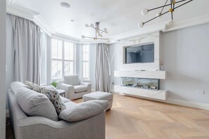 Un salon moderne avec un canapé gris, des fauteuils blancs, un grand téléviseur et un aquarium.