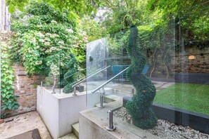 Un escalier extérieur en colimaçon, protégé par des parois de verre et doté de rampes en métal, est entouré d’une végétation luxuriante et d’un mur de briques.