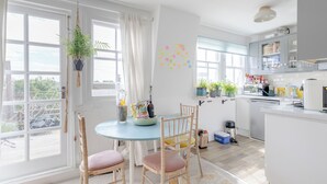 Valoisa keittiö, jossa on suuret ikkunat, ruokailutila pyöreän pöydän ja tuolien ympärillä sekä keittiötaso, jossa on kaappeja ja kodinkoneita.
