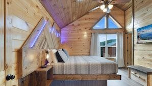 A wooden cabin bedroom with a loft bed, a ceiling fan, and a large window offering a view of the outdoors.