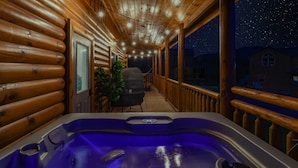 A wooden cabin with a hot tub on a porch, a chair, and a view of the night sky.