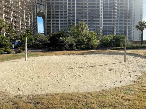 Una cancha de voleibol de arena, con una red, rodeada de vegetación y un edificio alto al fondo.