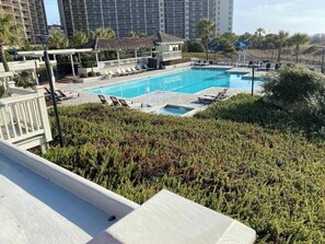 Una piscina con tumbonas y una zona de estar cubierta, rodeada de vegetación y altos edificios al fondo.