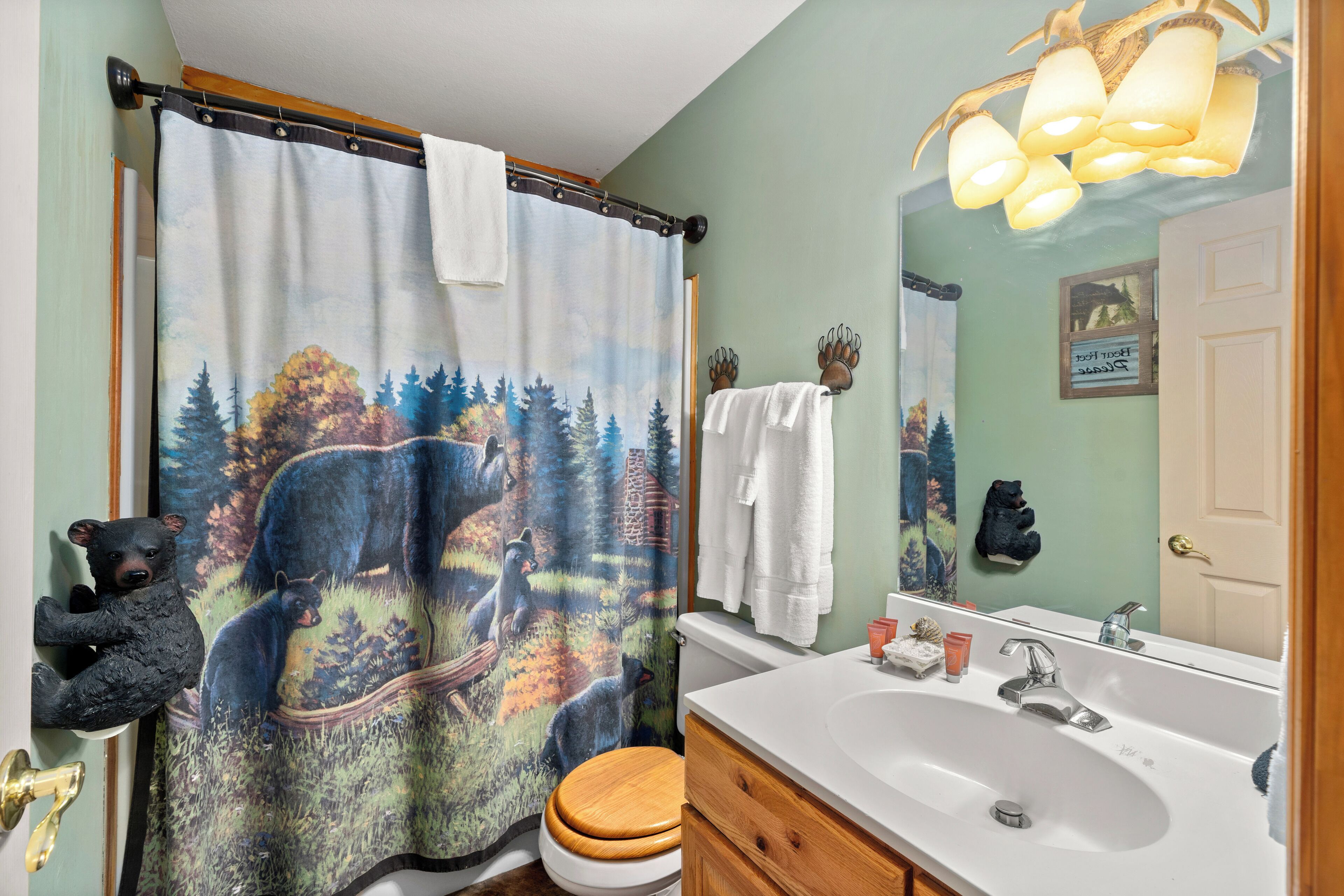 Ein Badezimmer mit Duschvorhang mit Bärenmotiv, einer Holzwc, einem weißen Waschbecken mit Toilettenartikeln und einem Spiegel, der den Raum reflektiert.