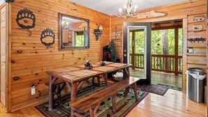 A wooden interior with a dining table, bench, chandelier, and a view through a door to an outdoor deck. - Creekfront Cabin w/ Hot Tub! Motorcycle Friendly! (Robbinsville)