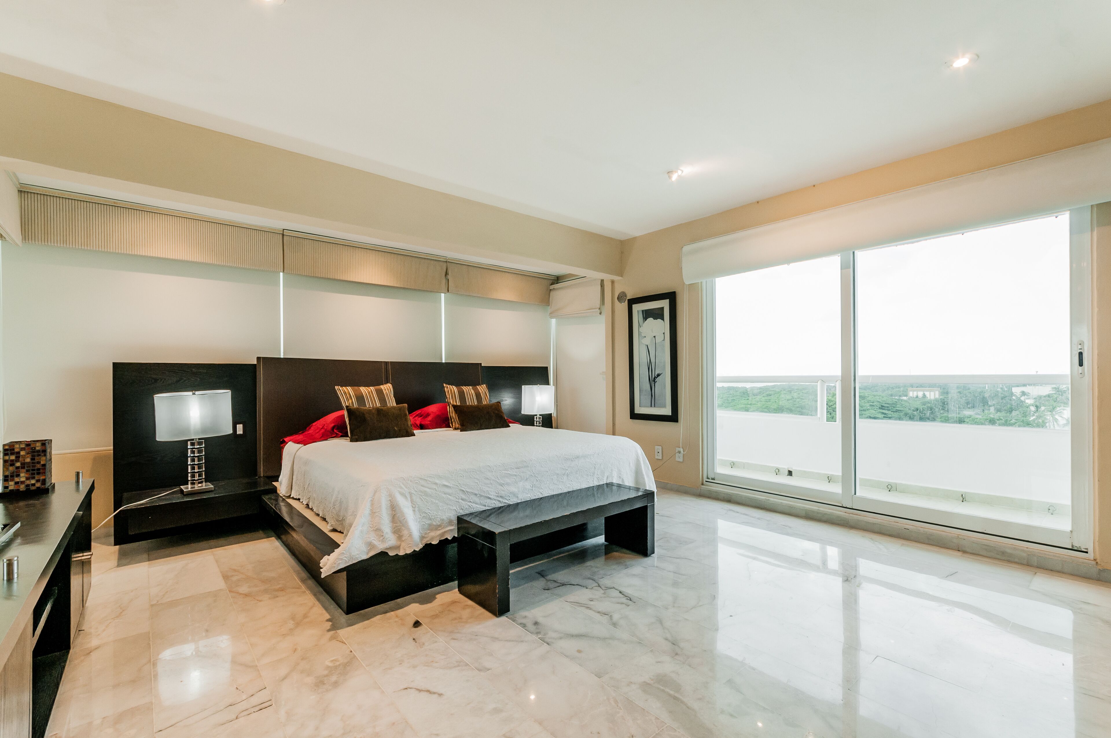A modern bedroom with a large bed, bedside lamps, a bench, and a view of the outdoors through large windows.