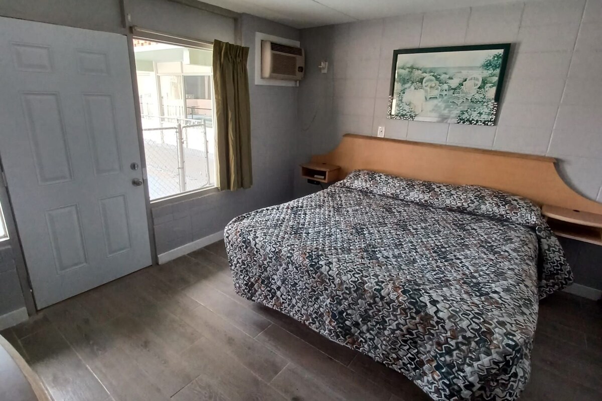 A hotel room with a bed, a nightstand, a window with curtains, an air conditioning unit, and a framed picture on the wall.