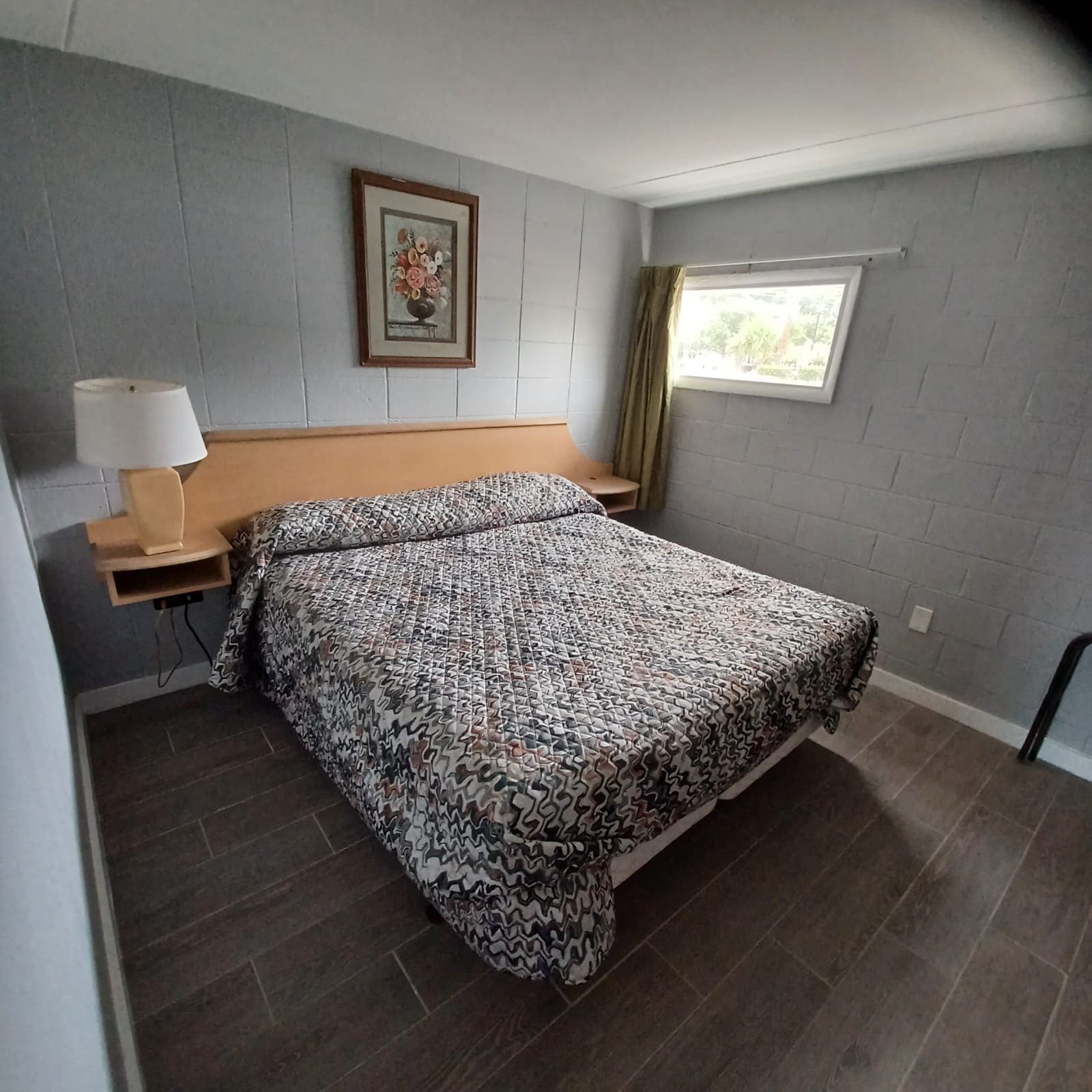 A bedroom with a bed, a nightstand, a lamp, a window with curtains, and a framed picture on the wall.