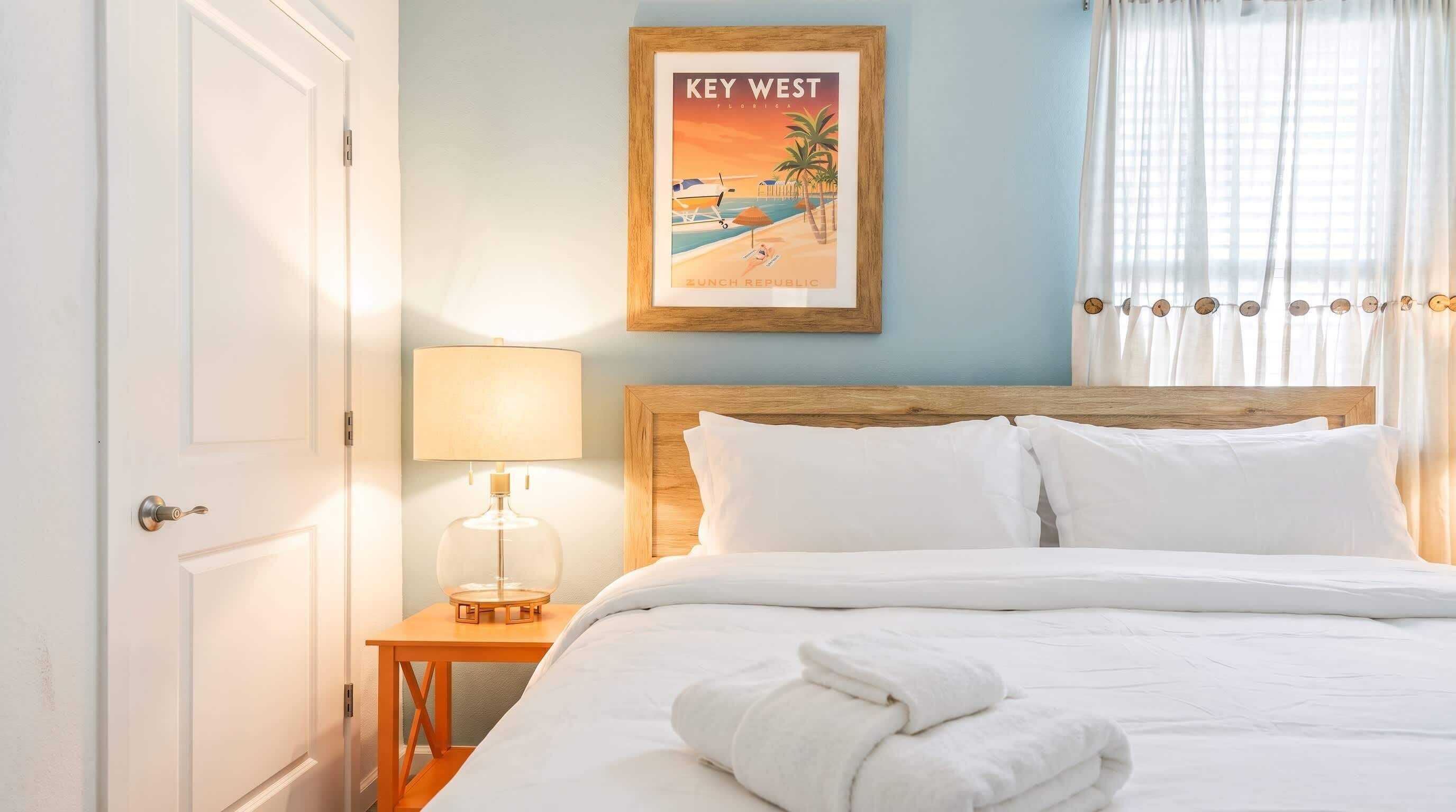 A hotel room with a bed, a nightstand with a lamp, a framed picture of Key West, and a door.