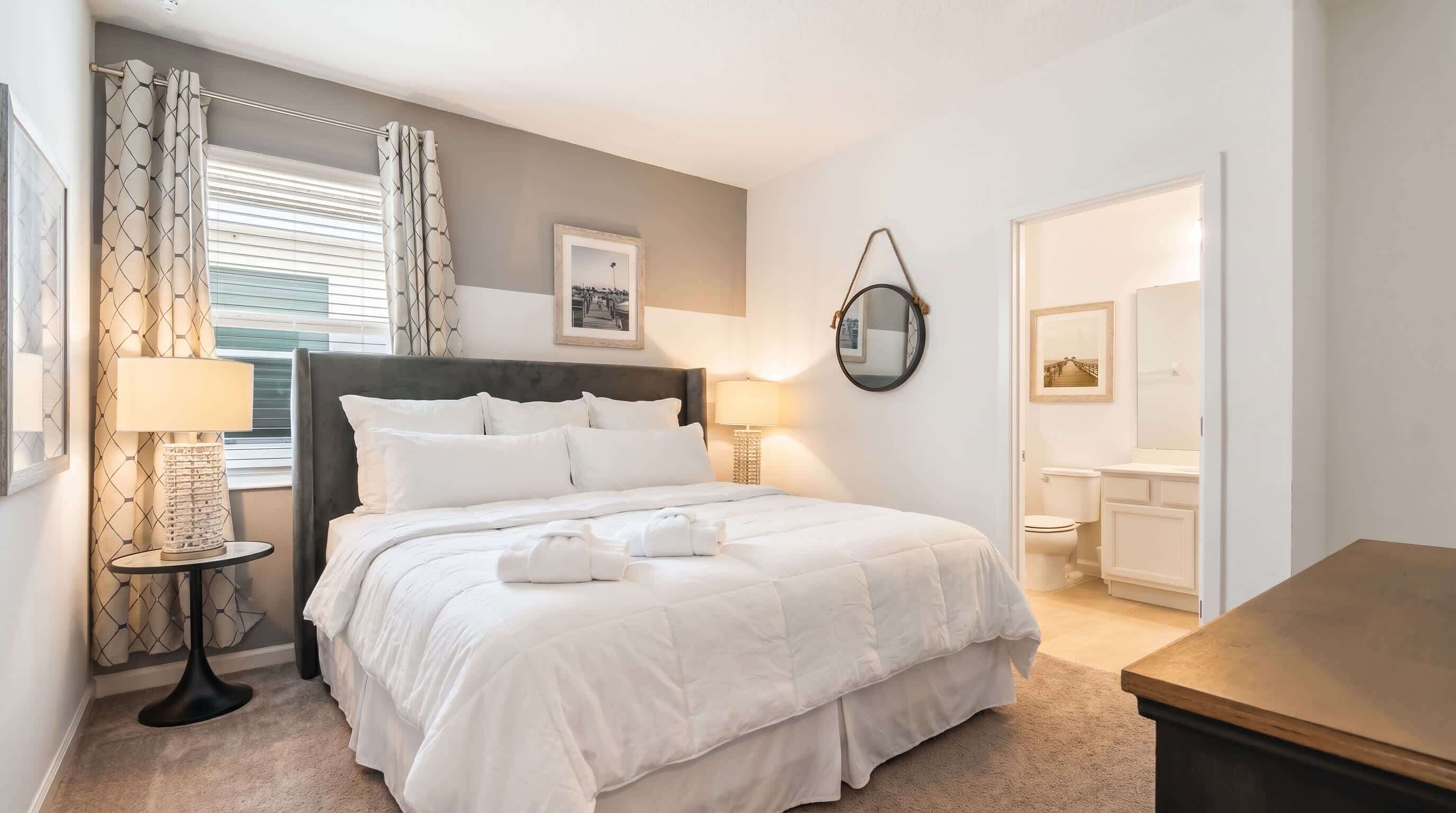 A hotel room with a bed, bedside lamps, a round mirror, a framed picture on the wall, and a bathroom visible through an open door.