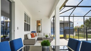 A modern outdoor patio with a glass table, blue chairs, a sofa with cushions, and a potted plant. - Spacious Family Reunion 15br Home Pool Spa Game Rm (Kissimmee)