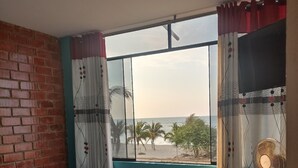 Ein Zimmer mit einem Bett, einem Schreibtisch, einem Fernseher und einem Fenster mit Blick auf Palmen und das Meer.