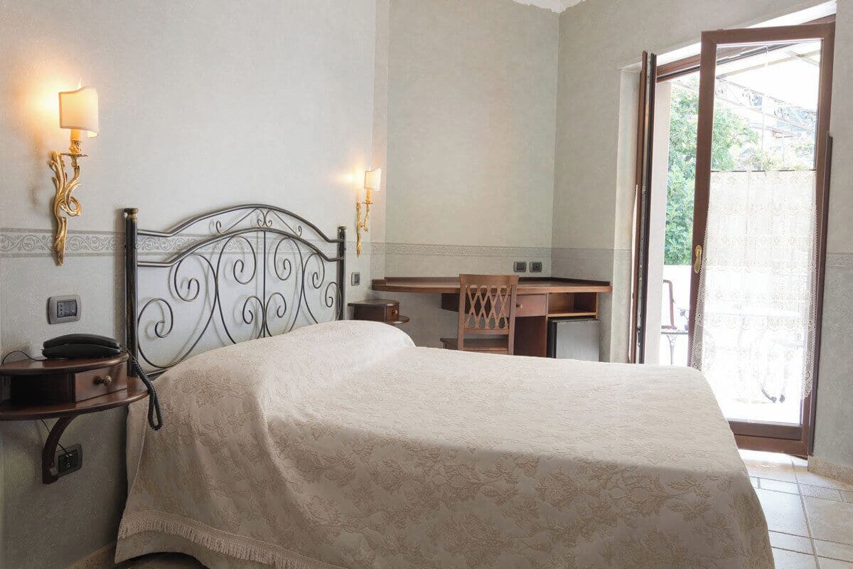 A bedroom with a bed, bedside table, lamp, desk, chair, and a view of the outdoors.