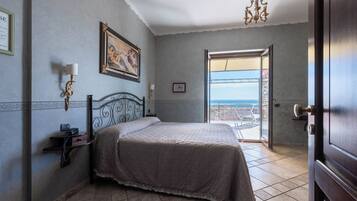 A bedroom with a bed, a bedside table with a lamp, a framed picture on the wall, and a view of the sea through an open door.