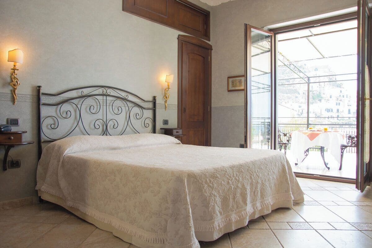 A bedroom with a bed, two wall-mounted lamps, a wooden door, and a balcony with a table and chairs.