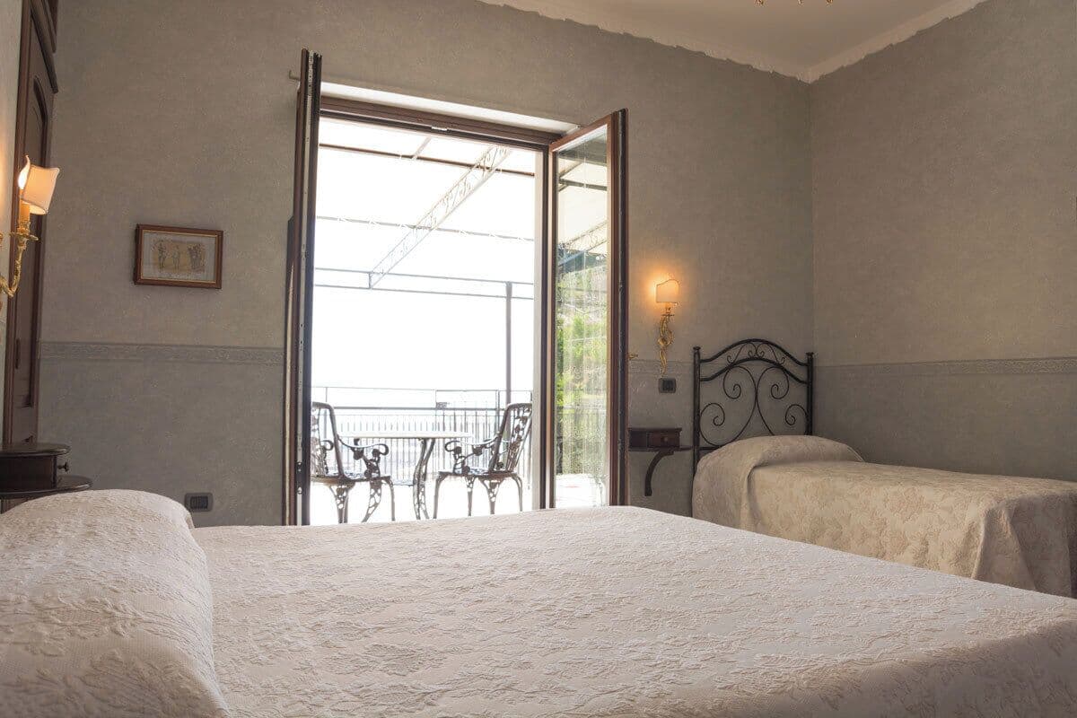 A bedroom with two beds, a balcony with chairs, and a view of the outside.