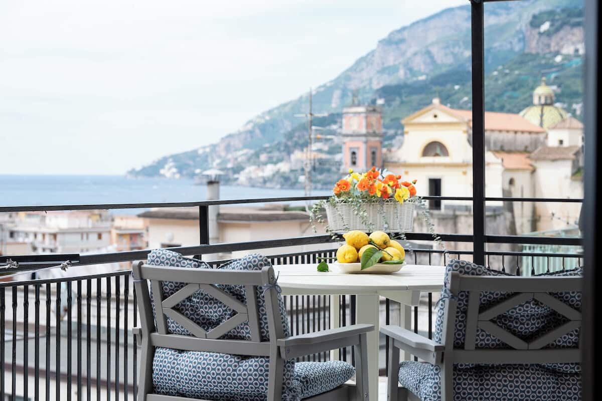 A balcony with a table set for two, featuring a vase with flowers and lemons, overlooking a coastal town with buildings and a mountain in the background.