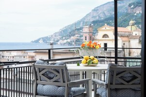 Un balcón con una mesa preparada para dos personas, con un florero con flores y limones, con vistas a un pueblo costero con edificios y una montaña al fondo.