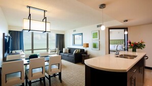 A hotel room with a dining area, a sofa, a TV, a large window, and a kitchen area.